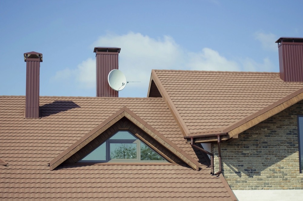 A brown roof with a gabled dormer window and two chimneys under a blue sky.